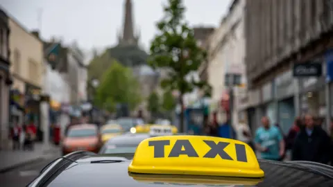 In the foreground the light on top of a taxi is seen. It has "TAXI" written in black capitals letters on a yellow background. Behind the taxi, but not in focus, a busy high street is visible with a church spire in the background