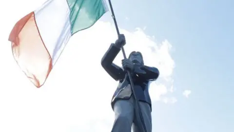 Close shot of a bronze Bobby Sands statue holding and orange white and green Irish flag against a blue sky.