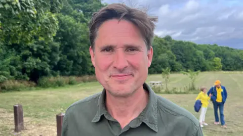 Charlie Maynard has short brown hair and is wearing a green shirt. Behind of him, out of focus, is a large grass field with a perimeter of bushes and trees.