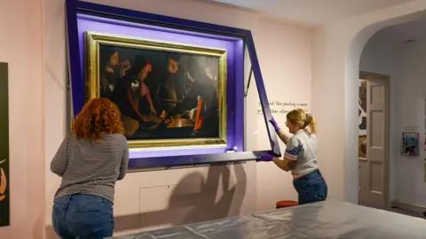 Stockton Borough Council The Dice Players is a painting that features a close-up scene of five people rolling dice on a table. It is quite dark but there is a splash of colour on some of their clothing, including orange and purple. They are wearing formal and elegant clothing. It is inside a large gold frame, which is being placed back into its case on a gallery wall at Preston Park Museum by two members of staff. The two women, pictured from behind, are closing the case.