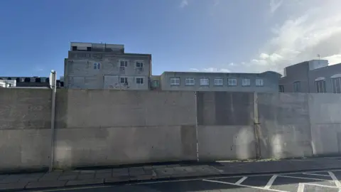 A boarded up wall with a few buildings behind it in vision and blue skies behind. A road is in front of the wall.