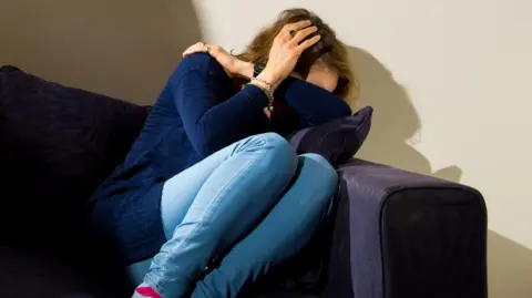 A woman in a blue jumper and light blue jeans who is cowering on a purple sofa. Her head is in her hands.