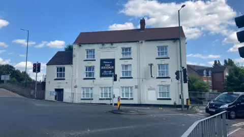 A white building with a brown roof sits by a road junction with traffic lights. A sign on the front reads: "Welcome to THE BRIDGE - Real Ales & Good Food."