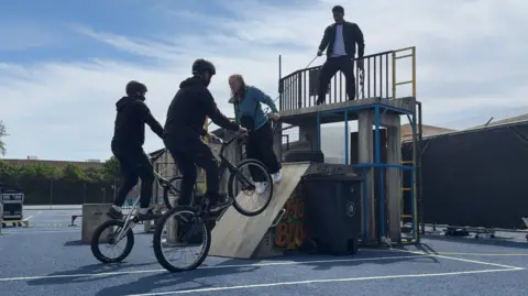 The photo shows a scene from a performance. Two men are dressed in black, wearing helmets and are on BMX bikes. They're doing a wheelie in front of a ramp. There is a woman with orange hair on the ramp, who is being pulled back by a rope round her torso by a man who is above her on a metal structure.