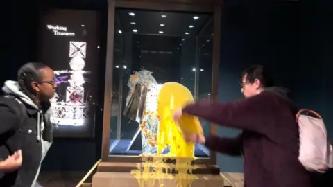 Two people stood next to the display case holding the Crown Jewels in the Tower of London, one is stood looking on while another is throwing yellow custard at the glass.