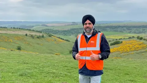 Bhupinder Sandhu A man wearing an orange high-vis vest with his thumbs up. He is stood at the top of a series of grassy hills