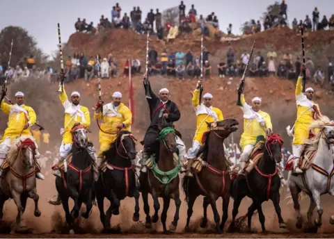 AFP Men lines up on horses. Most of them are wearing traditional Morocco clothes in yellow. They are raising rifles in the air. There are onlookers behind them.