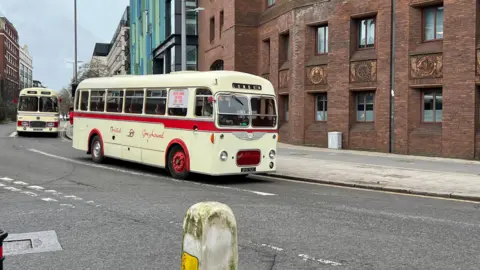 Two vintage coaches are parked on a street.  They are cream with red accents. A red brick building is visible in the background. 