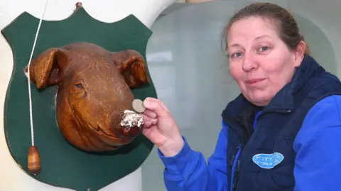 Kat Lister is standing beside a mounted wooden pig’s head sculpture attached to a shield-shaped green plaque on a wall. She holds a coin up to the pig’s snout, which has a small, worn or chipped patch on the surface. A pull cord with a wooden handle hangs next to the mounted sculpture on the left. Kat is wearing a blue long-sleeved top under a dark padded vest with a logo on the chest. The background shows part of a curved wall or doorway.