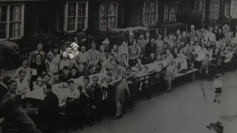 A black and white wide shot of a VE Day street party. Barry is standing wearing a white shirt and black tie, smiling into the camera.