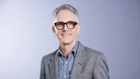 Falk Foundation A man with short grey hair is wearing black rimmed glasses, a grey suit jacket and a blue and white checked shirt