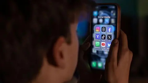 Tight close up of a teenager with his back to the camera, looking at a brightly lit smartphone screen with icons of social media ppas