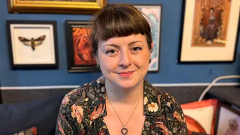 BBC A woman looking at the camera and smiling. She is sat in front of a blue wall which has a gallery of prints hung up on it.