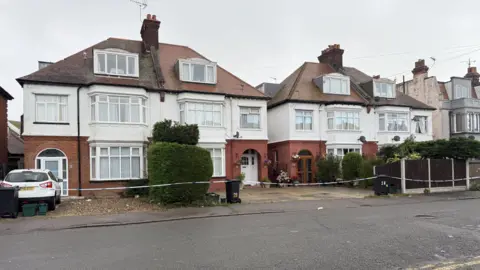 Elliot Deady/BBC Four semi-detached houses behind a police cordon. There is a grey cloudy sky.