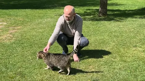 Property and estate manager Gavin Williamson petting the tabby cat Horatio on at Kelmscott Manor. It is a sunny day.