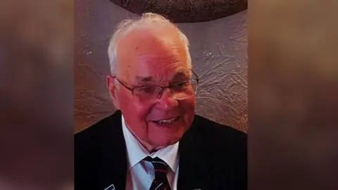 Sally Bluett Roy Mills in a dark suit, smiling. Image shows him as an older man with light-framed glasses.