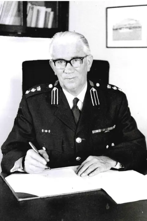 St John Ambulance and Rescue Service An older man with grey hair and glasses in an emergency services uniform. He sits at a desk ready to sign a book with a pen.