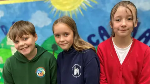 Three pupils smiling at the camera. There is a boy and two girls standing in front of a big mural with the sun on it and blue skies.