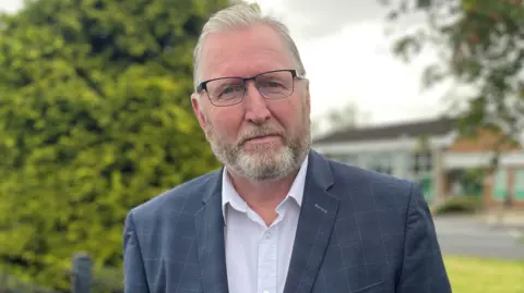 Doug Beattie has short white hair and a beard. He is smiling at the camera. He is wearing a pair of wire framed square glasses, a blue plaid suit jacket and a white collared shirt.