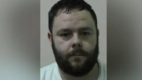 Mugshot of Binns. He has short dark hair and a thick dark beard and is wearing a white t-shirt.