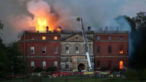 A red bricked stately home on fire. There are fire crews at the base. There is also two firefighters on an aerial platform firing water at the blaze which is coming from the roof on the left.