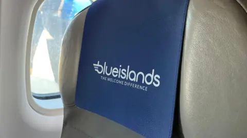 A view of a Blue Islands seat inside one of its aircrafts. The seat is positioned next to an oval shaped window. It is grey-ish in colour and made of leather. There is a blue seat cover which reads Blue Islands The Welcome Difference. 