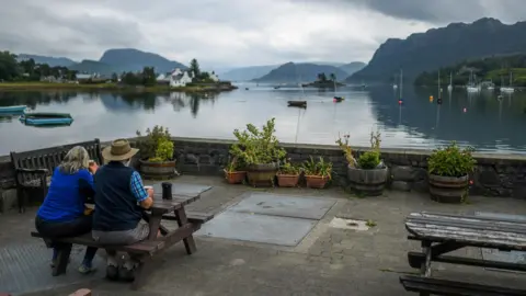 A woman and a man, who is wearing a wide brimmed hat, sit at a picnic bench drinking coffees and looking out over the bay at Plockton in Wester Ross.