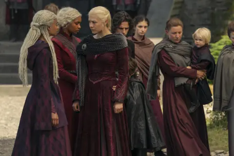 The cast of HBO's House of the Dragon includes six women, one holding a baby, all wearing floor-length burgundy dresses.