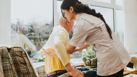 A young female carer helps an elderly male to get dressed by pulling on his jumper, as he sits in an armchair.