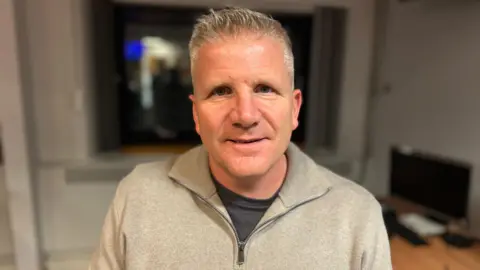 Chris Quinn - he is wearing a grey 1/4 zip jumper over a dark grey top. He has grey hair. The background is blurred but there is a wooden desk, a computer and a window in the background.