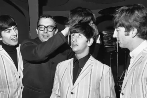 Getty Images Eric Morecambe is dressed in a Beatles-style coat and holding a Beatles-style wig above the head of Ringo Starr as George Harrison and John Lennon look on from either side.