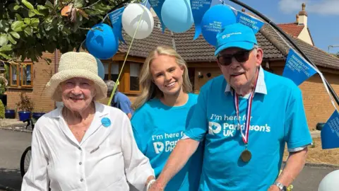 Imogen Treasure-Smith Doreen Treasure, wearing a white hat and shirt, holding the hand of David Treasure who is wearing a blue Parkinson's UK t shirt and cap and wearing a medal. Their granddaughter Imogen is behind them, wearing a blue Parkinson's UK t shirt with her blonde hair down. There is an arch behind them with Parkinson's UK balloons on. 