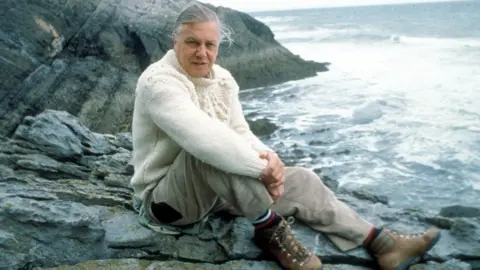David Attenborough sitting on a rocky shore on location for The Trials of Life