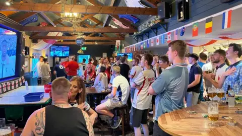 A group of people wearing England shirts are stood up in a bar lined with World Cup flags singing the national anthem.