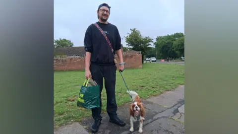 Liam Dunsdon-Stott-Bolton a man stood in all black, including a black cap and glasses. He is carrying a shopping bag in one hand and holding his dog's lead in the other hand. His dog is a small brown and white dog that is stood open mouthed with it's tongue out looking at the camera.