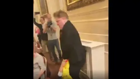 Andre Walker Andre Walker, dressed in a dark suit, is shown struggling to lift a man who is lying on the floor in an ornate room. A seated guest, a cameraman and another man can also be seen.