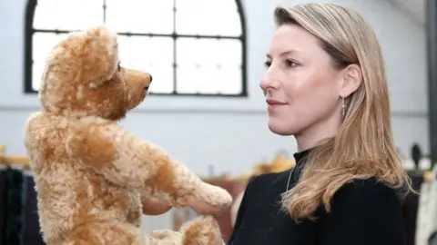 Merrythought A woman with blonde hair in a black top is smiling and holding up a golden coloured teddy hair