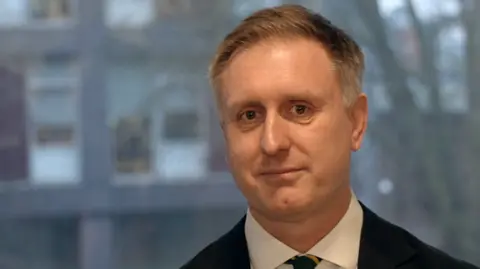 Vikki Irwin/BBC Ed Garrett is standing in front of a window, with an office building blurred in the background. He is wearing a dark suit and whit shirt with a green and yellow tie. he is smiling at the camera and the picture is a close up for him. 