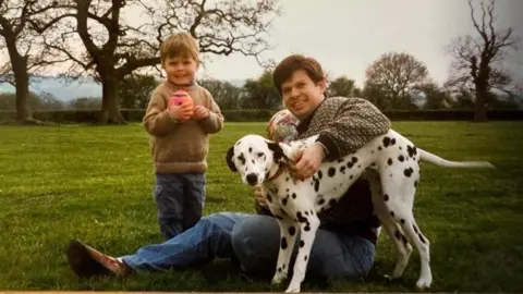 Family photo An archive picture of Dr Nicholas Bennett when his son Alexander was a boy. They are outside, in what looks like the countryside. Dr Bennet is sitting on the ground and playing with a Dalmatian. Alexander is holding a rugby ball and smiling for the camera.
