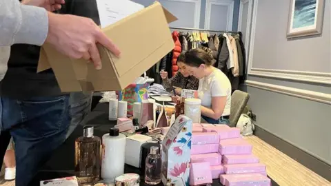 George Carden/BBC Volunteers pack cardboard boxes with products such as soap, shower gels for a winter hamper service. All the items are laid out on a table