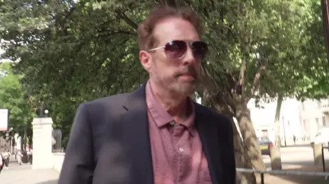 A man wearing a grey suit jacket and pink shirt walks along a tree-lined street. Only the top half of his torso and head are pictured. He has short brown hair and stubble and is wearing Aviator-style sun glasses.