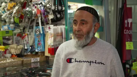 A man with a black knitted cap on his head, with grey facial hair and a long beard. He is wearing a grey jumper and is standing in front of a market stall which sells household goods