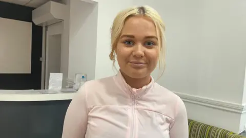 Picture of 17 year old college student Ella, who has bright blonde hair and is smiling towards the camera and wearing a zip up pink jacket. 