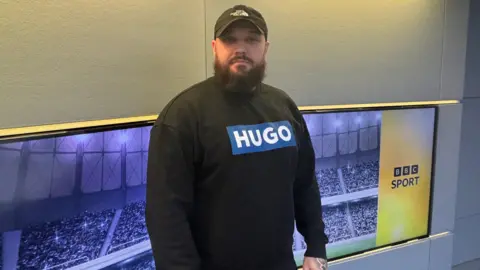 BBC A man with a hat, big beard and a black jumper which says "Hugo" stands in front of two screens with BBC Sport branding on the side.