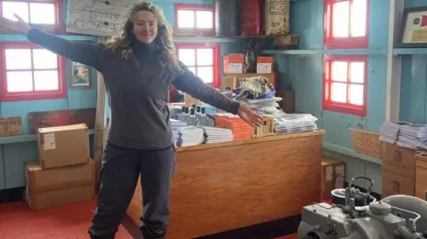 A picture of Dale Ellis. She has long hair and is stood smiling with her arms open wide in a room which has a desk in the background piled with items. 