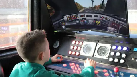 A pupil wearing a turquoise green jumper plays on a train simulator in the driver's cab of a Pacer train. 