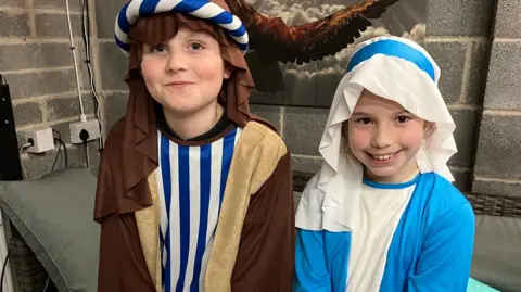 Oksi, who plays Joseph, and Elsie, who is Mary, are standing next to each other wearing traditional nativity costumes for the production. They are smiling at the camera. There is a cushion behind them and they are standing in a garage which has a picture on the wall.