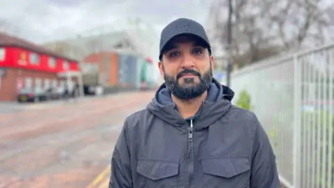 Man with a short beard and black cap stands outside of Old Trafford.