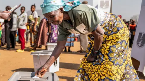 Reuters A woman wearing a blue headscarf and a yellow, patterned skirt bends over to place her ballot paper into a box.