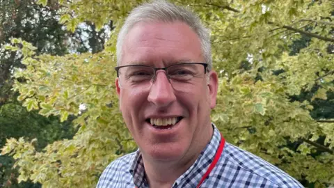 A man with short greying hair, wearing a blue and white shirt and a red lanyard, standing and smiling in front of a bush.